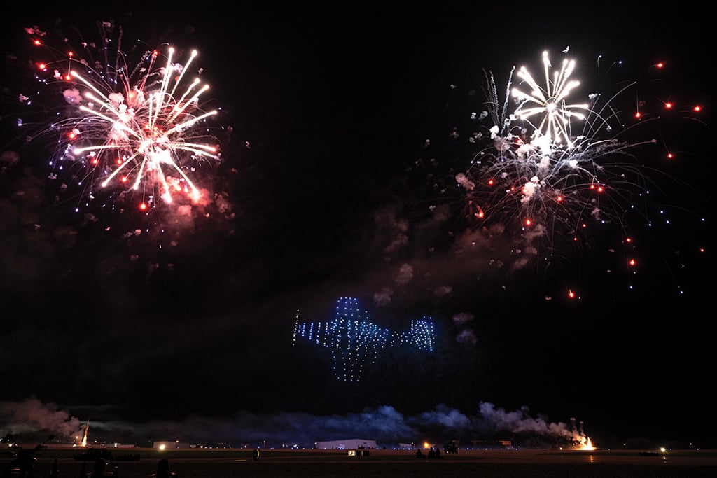 Red fireworks bursting, plus drones flying in the shape of an aircraft